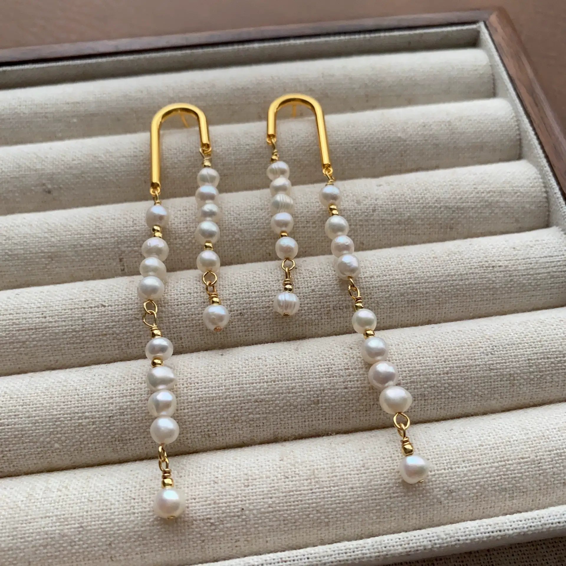 Asymmetric pearl tassel earrings with gold-tone arches displayed on a cream velvet jewelry tray.