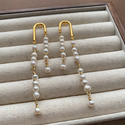 Asymmetric pearl tassel earrings with gold-tone arches displayed on a cream velvet jewelry tray.