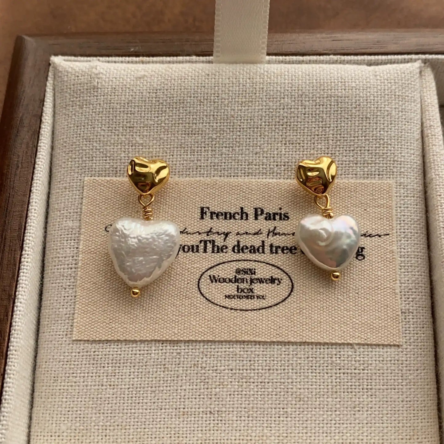 Heart baroque pearl earrings with gold-tone studs displayed on a beige linen jewelry card.