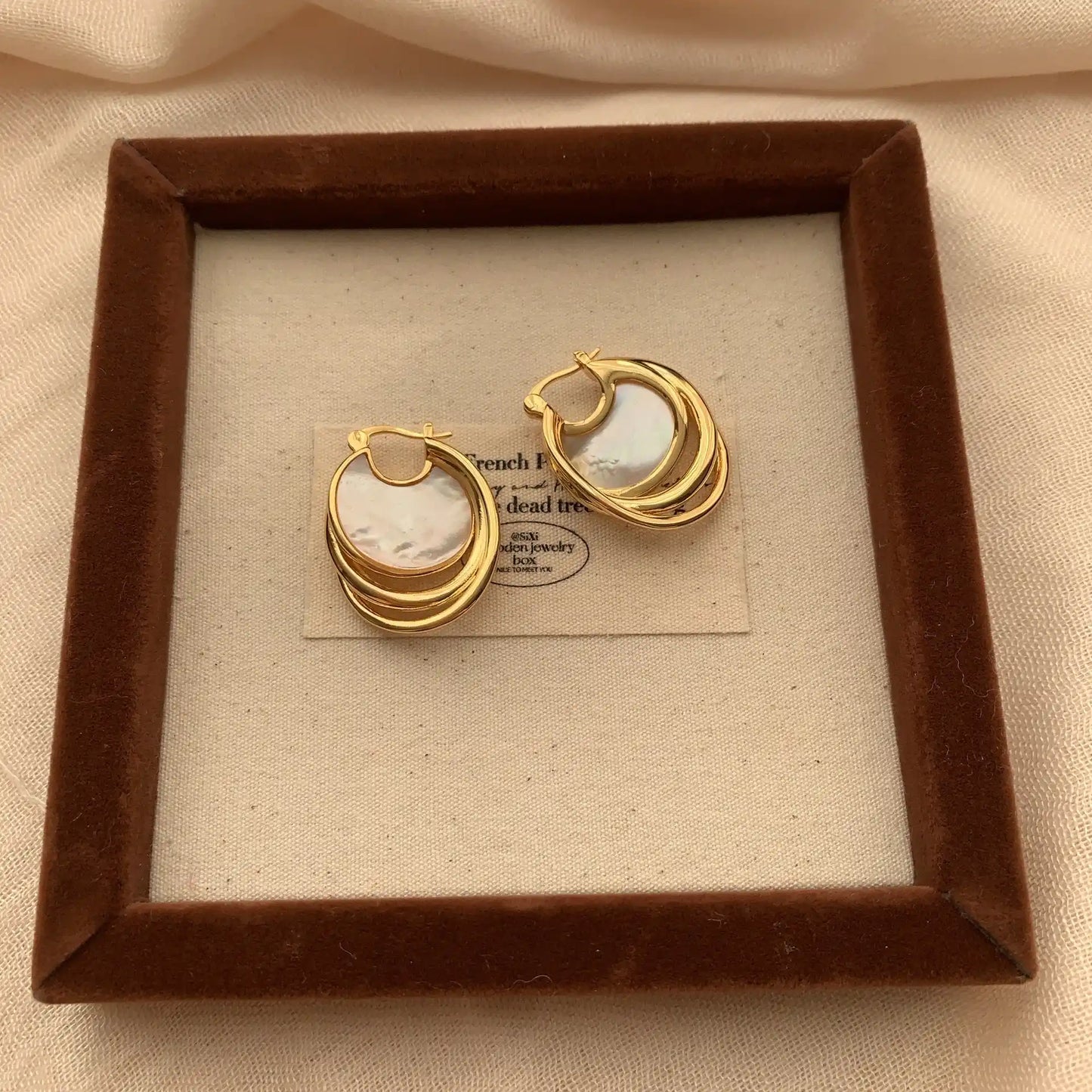 Circular mother of pearl earrings with sculptural gold hoops displayed on a framed fabric tray