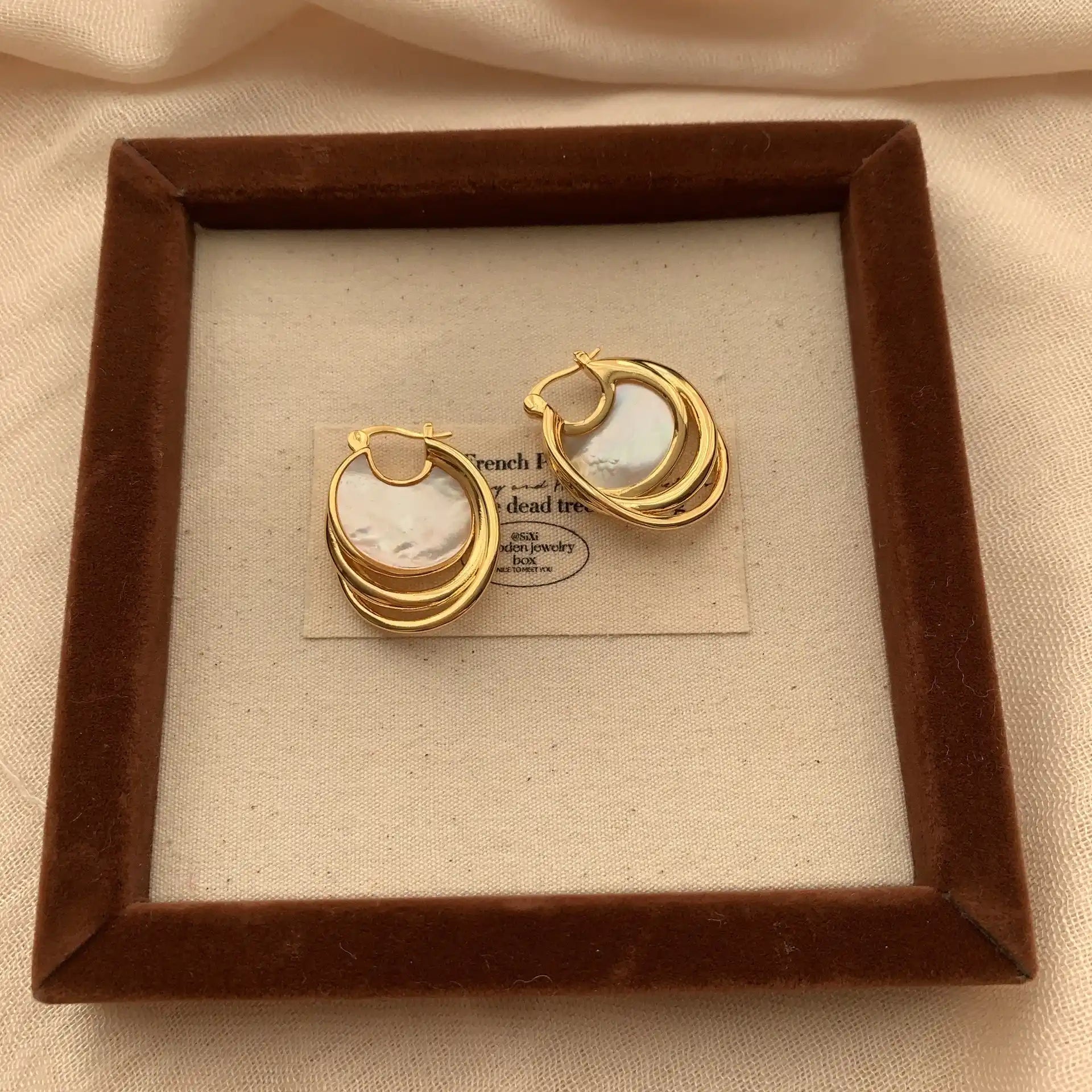 Circular mother of pearl earrings with sculptural gold hoops displayed on a framed fabric tray