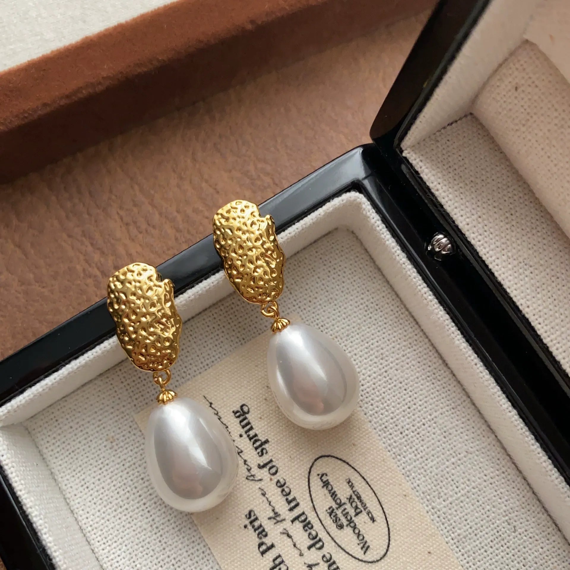 Teardrop pearl earrings with hammered gold-tone studs arranged in an open jewelry box.